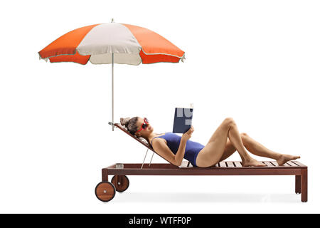 Profil de pleine longueur tourné d'une jeune femme dans un maillot de couché sur une chaise longue avec parapluie lire un livre isolé sur fond blanc Banque D'Images