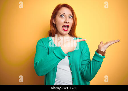 Belle jeune femme rousse portant chandail vert d'hiver isolé sur fond jaune étonné et souriant à la caméra tout en présentant à part Banque D'Images