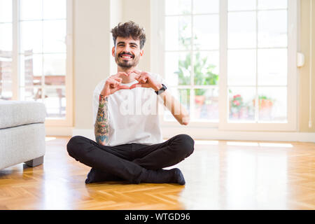 Jeune homme assis sur le travail occasionnel à la maison de plancher smiling in love montrant le symbole de coeur et la forme des mains. Concept romantique. Banque D'Images