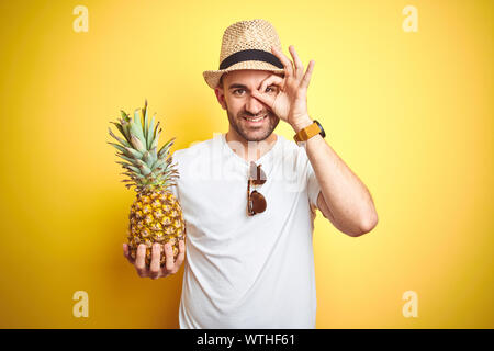 Jeune homme portant un chapeau d'été et la tenue sur fond jaune ananas avec happy smiling face ok faire signer avec la main sur l'œil à l'intermédiaire de finge Banque D'Images