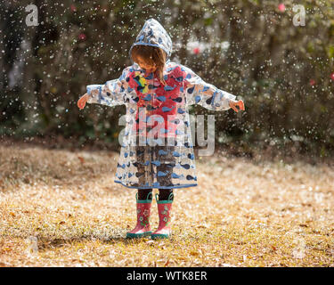 Petite fille dans l'imperméable et les bottes jouant dans la pluie. Banque D'Images