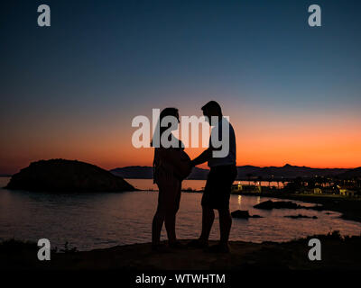 Silhouette of a pregnant couple sur une falaise avec le coucher du soleil et les lumières de la ville Banque D'Images