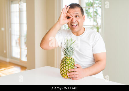 L'âge moyen de l'alimentation de l'homme à la maison de l'ananas frais tropicaux avec happy smiling face ok faire signe avec la main sur de l'oeil Banque D'Images