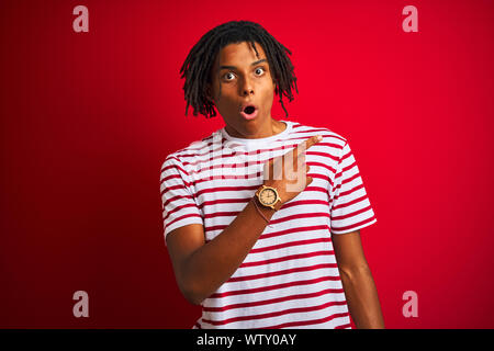 Jeune homme avec des dreadlocks afro portant des T-shirt à rayures sur fond rouge isolé permanent surpris pointant avec le doigt sur le côté, bouche ouverte am Banque D'Images
