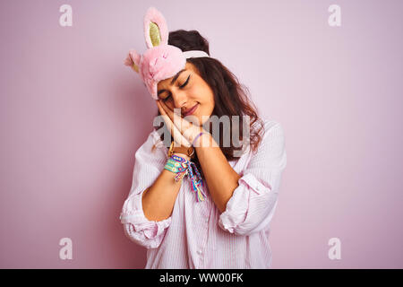 Belle Jeune femme portant un masque de sommeil et de pyjama sur fond rose isolé fatigué dormir rêver et posant avec les mains tout en souriant Banque D'Images