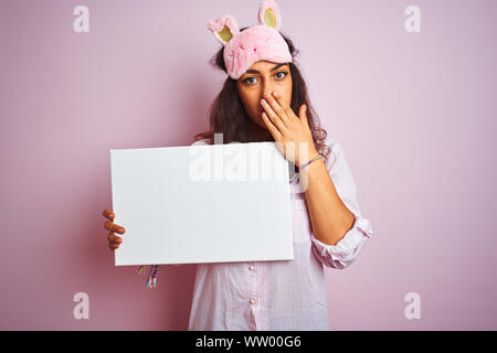 Jeune femme portant un masque de sommeil et de pyjama holding banner sur fond rose isolé couvrir la bouche à part choqué avec honte pour erreur, expressio Banque D'Images