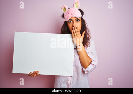 Jeune femme portant un masque de sommeil et de pyjama holding banner sur fond rose isolé couvrir la bouche à part choqué avec honte pour erreur, expressio Banque D'Images