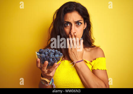 Young Beautiful woman holding bowl avec des bleuets debout sur fond jaune isolé couvrir la bouche à part choqué avec honte pour erreur, exp Banque D'Images