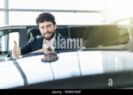 Cheerful car salesman smiling showing Thumbs up posant chez le concessionnaire beauté s'appuyant sur une nouvelle voiture à vendre Banque D'Images