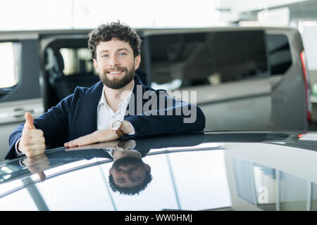 Cheerful car salesman smiling showing Thumbs up posant chez le concessionnaire beauté s'appuyant sur une nouvelle voiture à vendre Banque D'Images