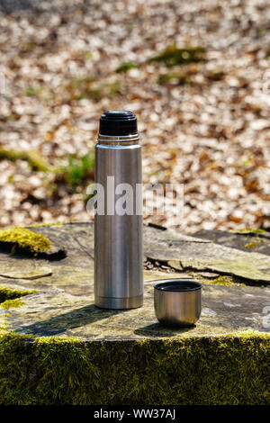 Bouteille thermos et une tasse de café ou de thé sur une souche d'arbre couverts de mousse dans la lumière du soleil dans les bois (vue de la hauteur des yeux, format vertical) Banque D'Images