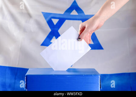 Jeune femme israélienne de mettre un bulletin dans l'urne le jour du scrutin. Close up of hand avec votes blancs sur fond du drapeau d'Israël. Immersive, de l'espace pour le texte. Banque D'Images