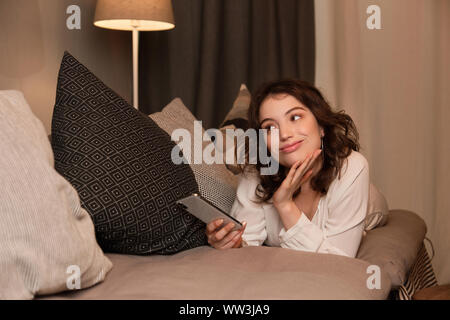 A curly girl se trouve sur un canapé et regarde rêveusement jusqu'en haut avec un téléphone dans sa main. Banque D'Images