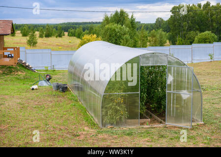 Une petite serre avec des plantations à l'intérieur est près de la maison, à côté d'elle cultive des plantes et l'herbe. Banque D'Images