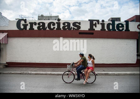 Un couple fait un tour en vélo le long des rues de la Havane, Cuba avec un panneau en hommage au regretté Fidel Castro Banque D'Images