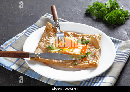 Close-up of Breton crêpes, galettes de sarrasin salées Bretons avec oeuf renversé, de fromage, de jambon sur une assiette blanche sur une table en béton Banque D'Images