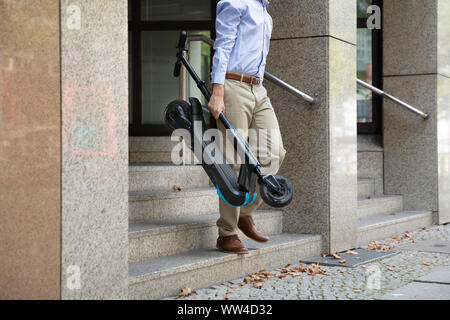 Homme portant l'E-scooter de travailler à la sortie de l'immeuble de bureaux Banque D'Images