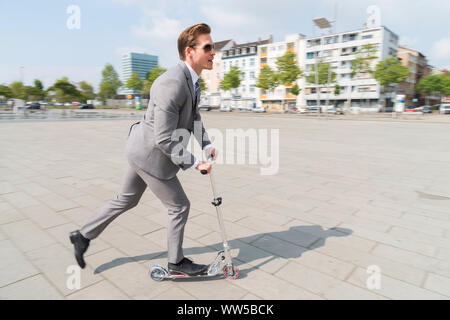 L'homme en costume gris riding scooter sur town square Banque D'Images