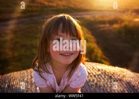 4-6 ans girl sitting on hay bale, souriant à la caméra, lumière du soir Banque D'Images