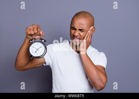 La photo en gros masculin sain peau noire il lui sa tête chauve macho perplexe tenir réveil en acier ne sais pas où couper la sonnerie wearing white t Banque D'Images