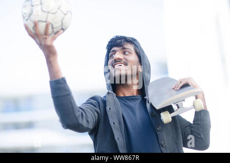 Jeune homme avec capuche à jouer au ballon et holding skateboard Banque D'Images
