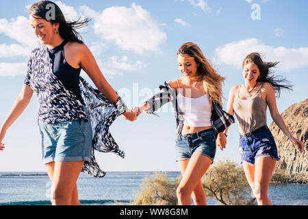 Trois jeunes jolies filles caucasiennes à marcher ensemble en tenant les mains chaque autres sourire et rire, joyeux notion de plaisir les femmes dans l'activité de loisirs de plein air près de l'océan et les montagnes Banque D'Images