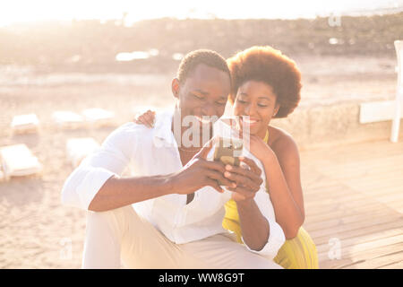 Young happy couple afro-américaine multi-ethnique de la race afro diversité raciale profiter de téléphone et internet dans l'activité de loisirs de plein air au cours de la brillante et coucher du soleil doré de lumière en arrière-plan, le sourire et rester ensemble Banque D'Images