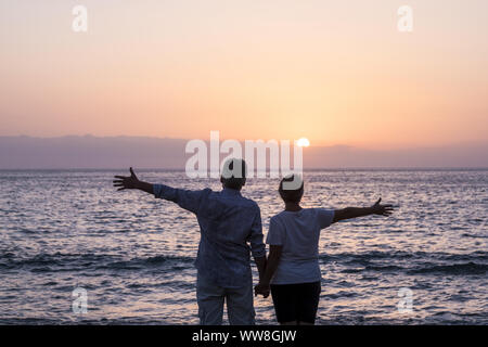 Concept de vacances, le tourisme, les voyages et les gens - happy senior couple sur une plage de galets de rire et plaisanter en regardant la mer au coucher du soleil avec les bras tendus, cheveux blancs