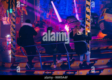 Les adolescents regarder les voitures sur une roue tournante fairground ride zip passé à une période nocturne de foire dans Hambourg, Allemagne Banque D'Images