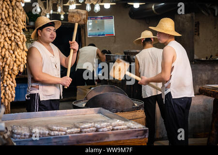 Xian, Chine - Juillet 2019 : Deux hommes de frapper fort avec l'aide de marteaux de bois de gros grains de crack qui sera utilisé dans la fabrication de sweet snack à vendre sur la str Banque D'Images