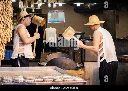 Xian, Chine - Juillet 2019 : Deux hommes de frapper fort avec l'aide de marteaux de bois de gros grains de crack qui sera utilisé dans la fabrication de sweet snack à vendre sur la str Banque D'Images