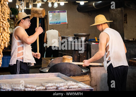 Xian, Chine - Juillet 2019 : Deux hommes de frapper fort avec l'aide de marteaux de bois de gros grains de crack qui sera utilisé dans la fabrication de sweet snack à vendre sur la str Banque D'Images