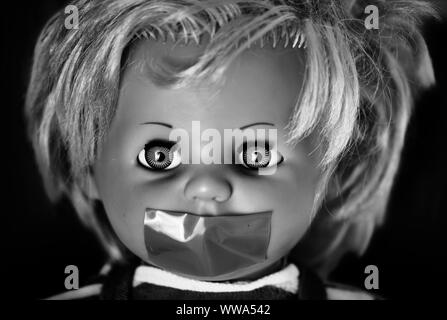 Tête de poupée Creepy en noir et blanc. Un visage effrayant d'une étrange poupée vintage avec sa bouche avec du ruban adhésif sur fond noir Banque D'Images