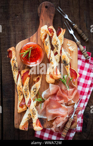 Batonnets de pâte feuilletée maison jambon fumé servi avec des tranches de speck italien et de romarin sur une planche à découper en bois rustique. Vie de frais généraux Banque D'Images