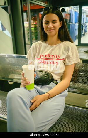 Portrait de fille avec des lunettes sur la tête posant assis sur un banc en centre commercial. Vêtus de T-shirt blanc, un pantalon bleu, noir à la taille. Banque D'Images