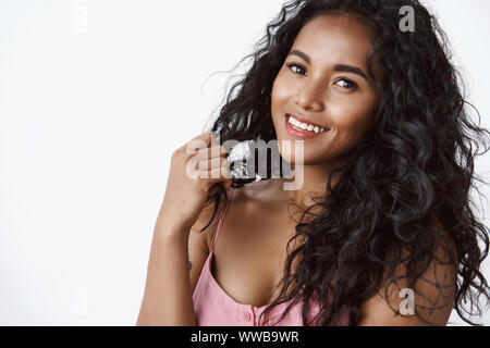 Close-up l'adjudication et féminine jeune femme afro-américaine avec curly hairstyle, jouant avec les boucles d'inclinaison et le regard caméra, sourire charmant que Banque D'Images