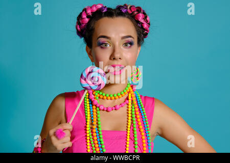 Jolie fille avec une coiffure tresses multicolores et lumineux maquillage, posant en studio sur un fond bleu, tenant une sucette à la main. Banque D'Images