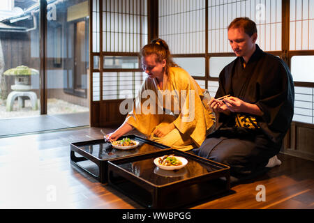 Couple en kimono assis seiza holding baguettes, mange la nourriture à la maison traditionnelle japonaise ryokan prix par table des assiettes en papier shoji portes coulissantes Banque D'Images