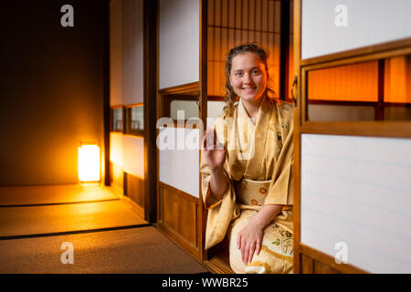 Maison traditionnelle japonaise ou ryokan avec gaijin caucasian woman en kimono et chaussettes tabi des signes avec la main de papier shoji ouvert les portes coulissantes assis o Banque D'Images