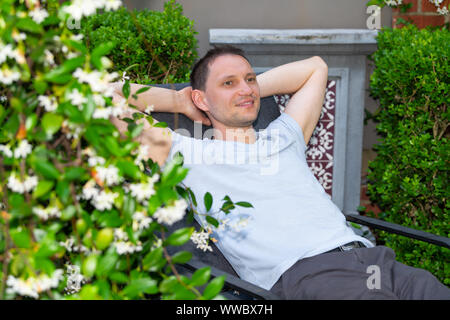 Young happy smiling man sur patio chaise longue avec les jambes jusqu'au printemps en plein air dans le jardin de fleurs jardin de plantes accueil Banque D'Images