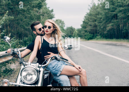 Jeune couple de bikers étroitement assis sur noir moto sur route près de la forêt verte Banque D'Images