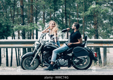 Jeune couple de bikers assis sur moto noir et l'homme de boire de l'alcool à partir de la bouteille sur la route près de la forêt verte Banque D'Images