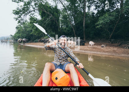 Young bearded Caucasian man paddling en kayak sur la rivière du Mékong avec le buffle d'eau sur le contexte, le Laos. Banque D'Images