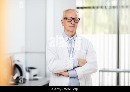 Portrait de friendly senior doctor wearing glasses looking at camera tout en se posant dans office standing with arms crossed, copy space Banque D'Images