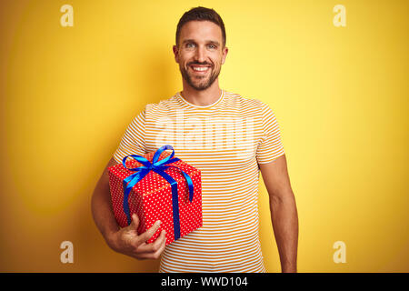 Jeune homme tenant un cadeau d'anniversaire sur jaune fond isolé avec un visage heureux et souriant debout avec un sourire confiant montrant les dents Banque D'Images