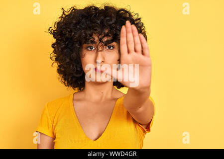 Jeune femme arabe avec des cheveux bouclés vêtus de t-shirt debout sur fond jaune isolé avec main ouverte faisant stop avec sérieux et confiance ex Banque D'Images