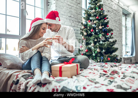 Beautiful couple in love, wearing santa hats, assis à côté d'un arbre de Noël joliment décoré et faire un toast avec des verres de champagne. Banque D'Images