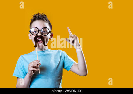 Un garçon dans une lumière t-shirt avec une moustache et des lunettes tient son index vers le haut contre un arrière-plan lumineux Banque D'Images