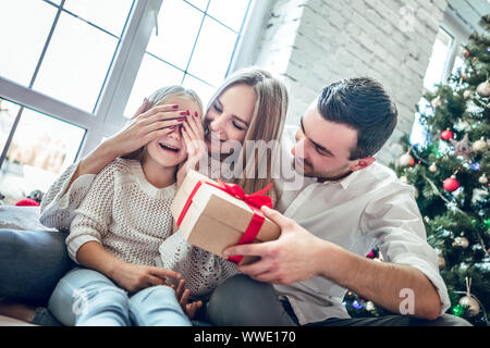 Portrait de jeune famille heureuse. Mère couvre ses yeux de fille en tant que père va faire surprise pour Fille donne à présent, près de l'arbre de Noël. Ha Banque D'Images