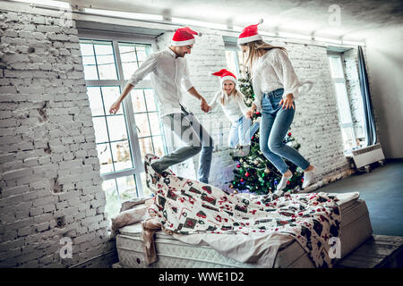 Moments drôles, young family jumping joyeusement sur le lit. Arrière-plan de l'arbre de Noël Banque D'Images